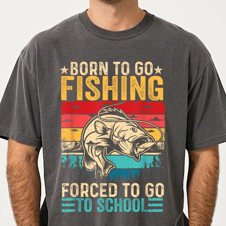 May include: Dark gray t-shirt with a colorful graphic. The design features a fish jumping out of the water, with the text "BORN TO GO FISHING" above and "FORCED TO GO TO SCHOOL" below. The graphic has a vintage, distressed look.