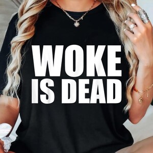 Op de afbeelding: Zwart T-shirt met de witte tekst "WOKE IS DEAD". Het T-shirt heeft een ronde hals. De afbeelding toont een close-up van het T-shirt.