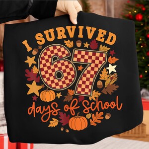 Op de afbeelding: Zwart T-shirt met de tekst "I Survived 67 days of school". De cijfers 6 en 7 hebben een rood-oranje ruitpatroon. Het ontwerp bevat pompoenen, bladeren en sterren in oranje en bruin.