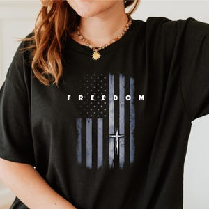 May include: Black t-shirt featuring a distressed American flag design with a cross and the word "FREEDOM" in white block letters. The flag design incorporates blue and white stars and stripes.