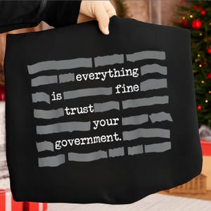 Op de afbeelding: Zwarte draagtas met de tekst "everything is fine trust your government" in wit en grijs. De tekst is gerangschikt in een blokstijl met horizontale grijze balken. De tas wordt door een persoon vastgehouden.