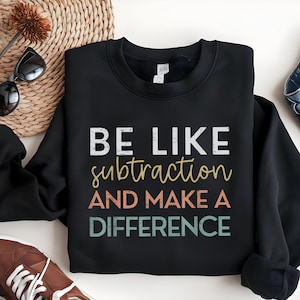 May include: Black sweatshirt with the text "BE LIKE subtraction AND MAKE A DIFFERENCE" in white, yellow, orange, and green. The sweatshirt is displayed on a white surface with sunglasses and a woven basket in the background.