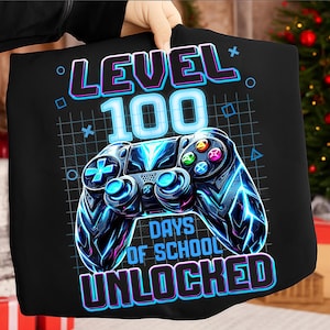 May include: Black t-shirt featuring a neon blue and teal video game controller design. The text reads "LEVEL 100 DAYS OF SCHOOL UNLOCKED" in a matching neon color scheme. The design includes geometric shapes.