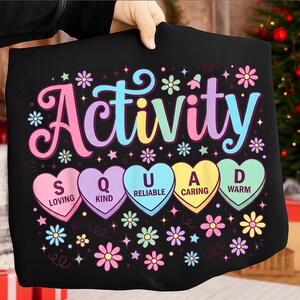Op de afbeelding: Zwart T-shirt met het woord "Activity" in pastelkleuren en het woord "Squad" uitgespeld in hartvormige snoepjes. De harten hebben woorden als "Loving" en "Kind". Het shirt is versierd met bloemen en sterren.