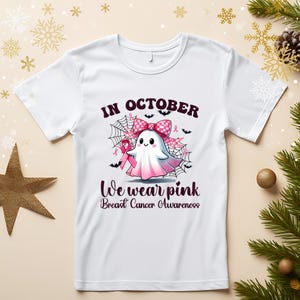 Op de afbeelding: Wit T-shirt met een afbeelding van een spook met een roze strik en lint, met de tekst "In October We Wear Pink Breast Cancer Awareness". Het ontwerp bevat spinnenwebben en vleermuizen.
