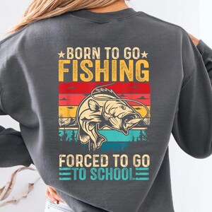 May include: Dark gray sweatshirt with a graphic that reads "BORN TO GO FISHING" above a fish illustration and "FORCED TO GO TO SCHOOL" below. The design features a fish illustration with a colorful sunset background.