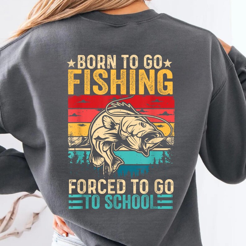 May include: Dark gray sweatshirt with a graphic that reads "BORN TO GO FISHING" above a fish illustration and "FORCED TO GO TO SCHOOL" below. The design features a fish illustration with a colorful sunset background.