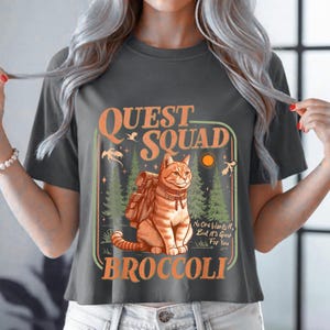 Könnte beinhalten: Dunkelgraues T-Shirt mit einer Grafik, die eine orangefarbene Tabby-Katze mit Rucksack zeigt, mit den Worten "QUEST SQUAD BROCCOLI". Das Design umfasst Bäume, Vögel und den Text "No One Wants It, But It's Good For You."
