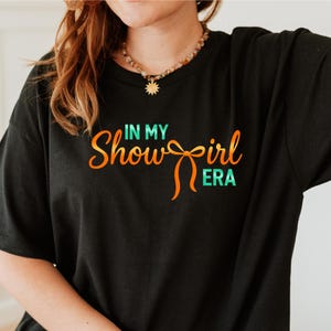 Op de afbeelding: Zwart T-shirt met de tekst "IN MY Showgirl ERA" in oranje en teal. Het shirt heeft een relaxte pasvorm en korte mouwen. De tekst is in een speels, gestileerd lettertype.