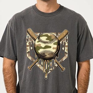 May include: Dark grey t-shirt with a baseball design. The baseball has a camouflage pattern and is dripping. Two crossed baseball bats and a home plate shape complete the design. A casual wear item.
