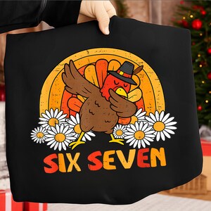 Op de afbeelding: Zwart T-shirt met een cartoon kalkoen die een dab doet, een pelgrimshoed draagt en tegen een regenboogachtergrond staat. Witte madeliefjes staan onderaan en de tekst "SIX SEVEN" staat in oranje en geel.