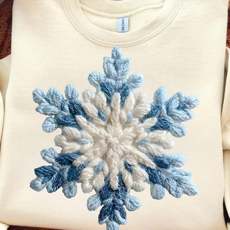 May include: Cream-coloured sweatshirt with a large, embroidered snowflake design. The snowflake is made with varying shades of blue and white thread, creating a textured, three-dimensional effect. The sweatshirt has a classic crew neck and long sleeves.