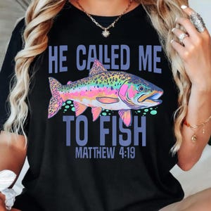 May include: Black t-shirt featuring the text "HE CALLED ME TO FISH" and "MATTHEW 4:19" in blue. A vibrant illustration of a rainbow trout, with pink, yellow, green, and blue colours, is the central design element.