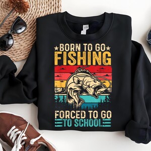 May include: Black sweatshirt with a graphic that reads "BORN TO GO FISHING" above a fish illustration and "FORCED TO GO TO SCHOOL" below. The design features a sunset color scheme with a fish and trees.