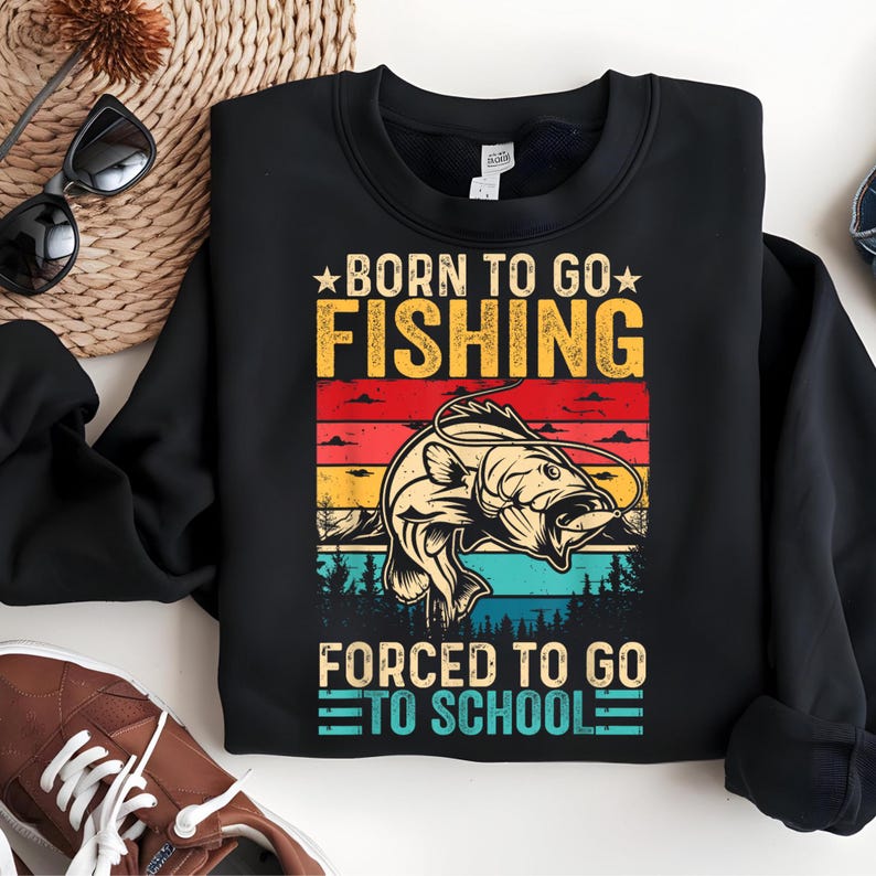 May include: Black sweatshirt with a graphic that reads "BORN TO GO FISHING" above a fish illustration and "FORCED TO GO TO SCHOOL" below. The design features a sunset color scheme with a fish and trees.