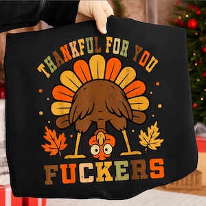 Op de afbeelding: Zwart T-shirt met een Thanksgiving-thema grafisch ontwerp. Het ontwerp toont een cartoon kalkoen ondersteboven met de tekst "THANKFUL FOR YOU FUCKERS" in oranje en geel. Herfstbladeren en stippen maken het ontwerp compleet.