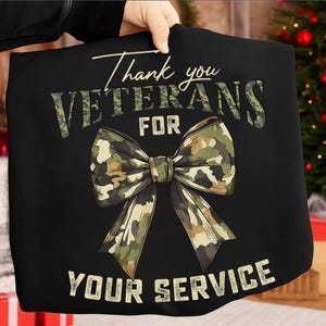 Op de afbeelding: Zwart T-shirt met de tekst "Thank you VETERANS for YOUR SERVICE" in witte en camouflage letters. Een grote camouflage strik is gecentreerd onder het woord "FOR". Het T-shirt wordt door een persoon vastgehouden.