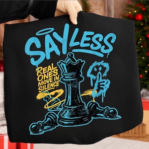 Op de afbeelding: Zwart T-shirt met een schaakthema. Het ontwerp bevat een koningin en pionnen, met de woorden "SAY LESS" in blauwe graffiti-stijl. De tekst "REAL ONES MOVE IN SILENCE" is geel. Een blauw handgebaar is ook inbegrepen.