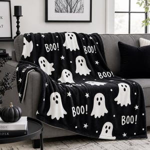 Op de afbeelding: Een zwarte plaid met een herhalend patroon van witte spoken, kleine witte sterren en de tekst "BOO!" in het wit gedrukt. De deken heeft een zachte, pluche textuur en is gedrapeerd over een grijze bank, wat zorgt voor een contrastrijk ontwerp.