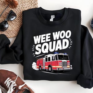 Può includere: Felpa nera con la scritta "WEE WOO SQUAD" sopra una grafica di un camion dei pompieri rosso e bianco. L'autopompa ha luci blu ed emette fumo. La felpa è uno stile girocollo.