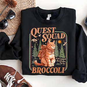 May include: Black sweatshirt with a graphic featuring an orange tabby cat wearing a backpack, with the words "QUEST SQUAD" and "BROCCOLI". The design includes trees, a sun, and the text "No One Wants It, But It's Good For You."