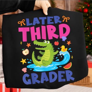 Könnte beinhalten: Schwarzes T-Shirt mit dem bunten Schriftzug "Later Third Grader". Das Design zeigt ein grünes Alligator, eine gelbe Ente, Sterne, Äpfel und eine Wassermelone.