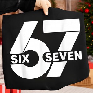 Op de afbeelding: Zwart T-shirt met het nummer "67" in grote witte blokletters. Het woord "SIX" staat onder de "6" en het woord "SEVEN" onder de "7". Het T-shirt wordt door een persoon vastgehouden.