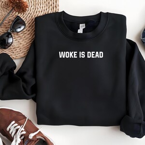 Op de afbeelding: Zwart sweatshirt met witte tekst "WOKE IS DEAD". Het sweatshirt wordt getoond op een neutrale ondergrond met een zonnebril, een geweven placemat en bruine sneakers.