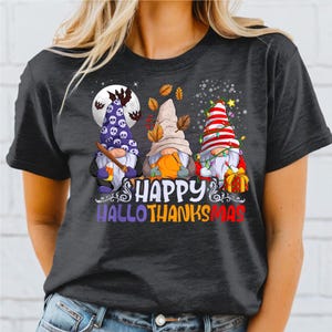 Op de afbeelding: Donkergrijs t-shirt met drie kabouters met feestelijke hoeden. De kabouters zijn versierd met Halloween- en Thanksgiving-thema's. De tekst "HAPPY HALLOTHANKSMAS" staat eronder.