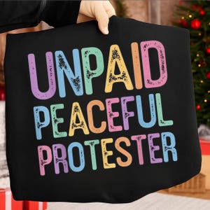 Op de afbeelding: Zwart T-shirt met de tekst "UNPAID PEACEFUL PROTESTER" in grote, kleurrijke, vervaagde letters. De tekst is in een regenboog van pastelkleuren, waaronder paars, blauw, geel en roze. Het shirt wordt door een persoon omhoog gehouden.