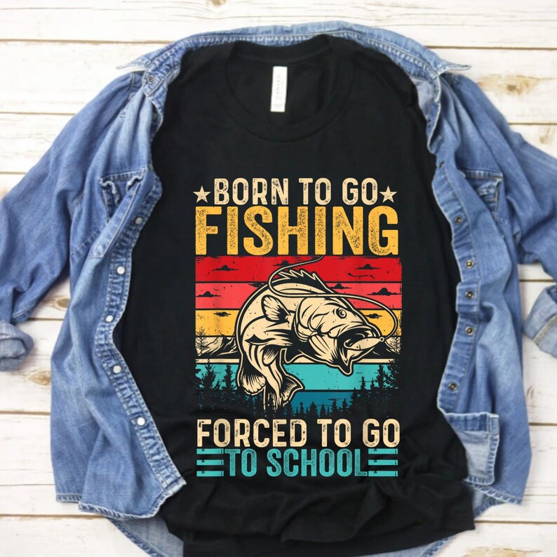 May include: Black t-shirt with a colorful graphic design. The design features a fish illustration and the text "BORN TO GO FISHING" above "FORCED TO GO TO SCHOOL". The shirt is displayed on a wooden surface with a denim jacket.