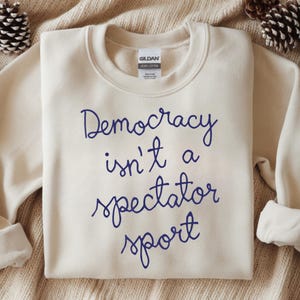 Può includere: Felpa color crema con la scritta "Democracy isn't a spectator sport" in blu navy. L'etichetta del marchio Gildan è visibile. La felpa è esposta su una superficie strutturata.