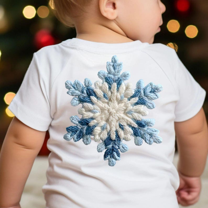 May include: A white t-shirt with short sleeves, adorned with a large, embroidered snowflake. The snowflake design uses white and various shades of blue. The t-shirt is designed for a child, perfect for winter wear.