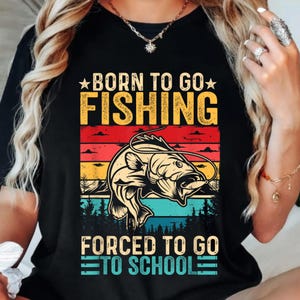 May include: Black t-shirt with a fishing-themed graphic. The design features a fish with a fishing line, set against a sunset backdrop of red, yellow, and blue stripes. Text reads "BORN TO GO FISHING" and "FORCED TO GO TO SCHOOL."
