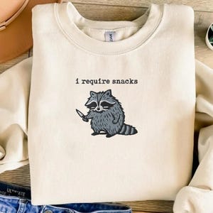 May include: Cream-colored sweatshirt with the text "I require snacks" above a cartoon raccoon holding a knife. The raccoon is gray and black with a fluffy tail. The sweatshirt is folded.