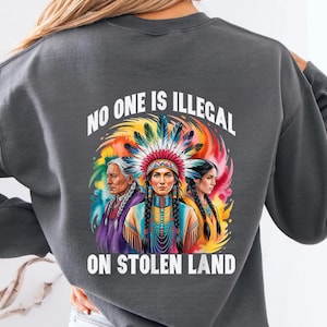 Op de afbeelding: Donkergrijze sweatshirt met een levendige afbeelding van drie inheemse Amerikaanse vrouwen en de tekst "NO ONE IS ILLEGAL ON STOLEN LAND". Het ontwerp bevat een kleurrijke hoofdtooi en een regenboogachtergrond.