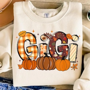 Op de afbeelding: Crèmekleurige sweatshirt met het woord "GIGI" in een herfstthema. De letters zijn versierd met ruit-, bloemen- en bladerpatronen en worden vergezeld door drie pompoenen. Perfect voor de herfst.