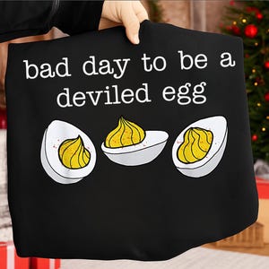 Op de afbeelding: Zwarte draagtas met de witte tekst "bad day to be a deviled egg". Het ontwerp toont drie gevulde eieren met gele vulling en een snufje paprika.
