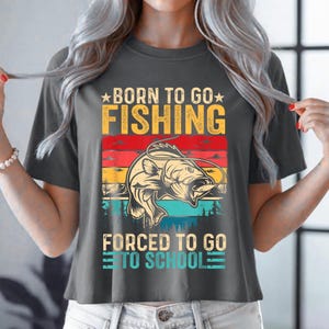 May include: Dark gray t-shirt with a graphic that reads "BORN TO GO FISHING" above a fish illustration and "FORCED TO GO TO SCHOOL" below. The graphic features a fish jumping out of the water with a colorful sunset background.