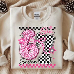 Könnte beinhalten: Cremefarbenes Sweatshirt mit rosa und schwarzer Grafik. Die Grafik zeigt die Zahlen "6" und "7" mit Leopardenmuster bzw. Schachbrettmuster. Die Wörter "Six" und "Seven" sind ebenfalls enthalten.
