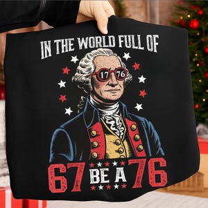 Op de afbeelding: Zwart T-shirt met een afbeelding van George Washington met een rood montuur bril met "1776". De tekst luidt "IN THE WORLD FULL OF" boven de afbeelding en "67 BE A 76" eronder, met rode en witte sterren.