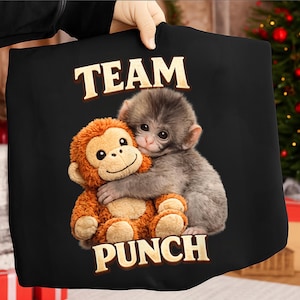 Op de afbeelding: Zwarte draagtas met de woorden "TEAM PUNCH" in gouden letters. De tas heeft een oranje pluchen aap die een grijze aap omhelst. De afbeelding staat tegen een vage achtergrond.