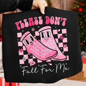 Op de afbeelding: Zwart T-shirt met een afbeelding van twee roze cartoon sokken met gezichten, tegen een roze en witte geblokte achtergrond. De tekst "PLEASE DON'T" staat boven de sokken en "Fall For Me" staat eronder.
