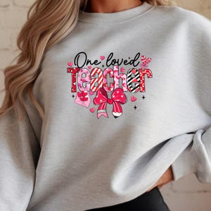 Op de afbeelding: Een lichtgrijs sweatshirt met de tekst "One loved Teacher" in een kleurrijk ontwerp. Het woord "Teacher" is in rode, witte en zwarte patronen, met een grote roze strik en hartjes. Een leuk cadeau voor een leraar.