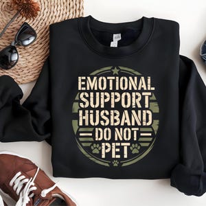 Op de afbeelding: Zwart sweatshirt met de tekst "EMOTIONAL SUPPORT HUSBAND - DO NOT PET" in een cirkelvormig ontwerp. Het ontwerp bevat pootafdrukken en een ster. Het sweatshirt wordt gepresenteerd op een witte ondergrond.