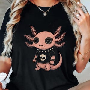 Op de afbeelding: Zwart T-shirt met een cartoon axolotl in punkstijl. De roze axolotl draagt een zwart shirt met een schedelmotief en een kraag met studs. De axolotl heeft grote ogen en een ietwat chagrijnige uitdrukking.