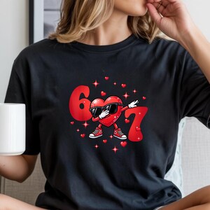 May include: Black t-shirt featuring a red heart character wearing sunglasses and sneakers, dabbing with the numbers 6 and 7 in red. Small red hearts and stars surround the heart character.