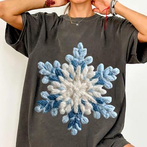 May include: Dark grey t-shirt with a large, textured snowflake design. The snowflake is made with white and blue yarn, creating a 3D effect. The t-shirt has a relaxed fit and short sleeves.