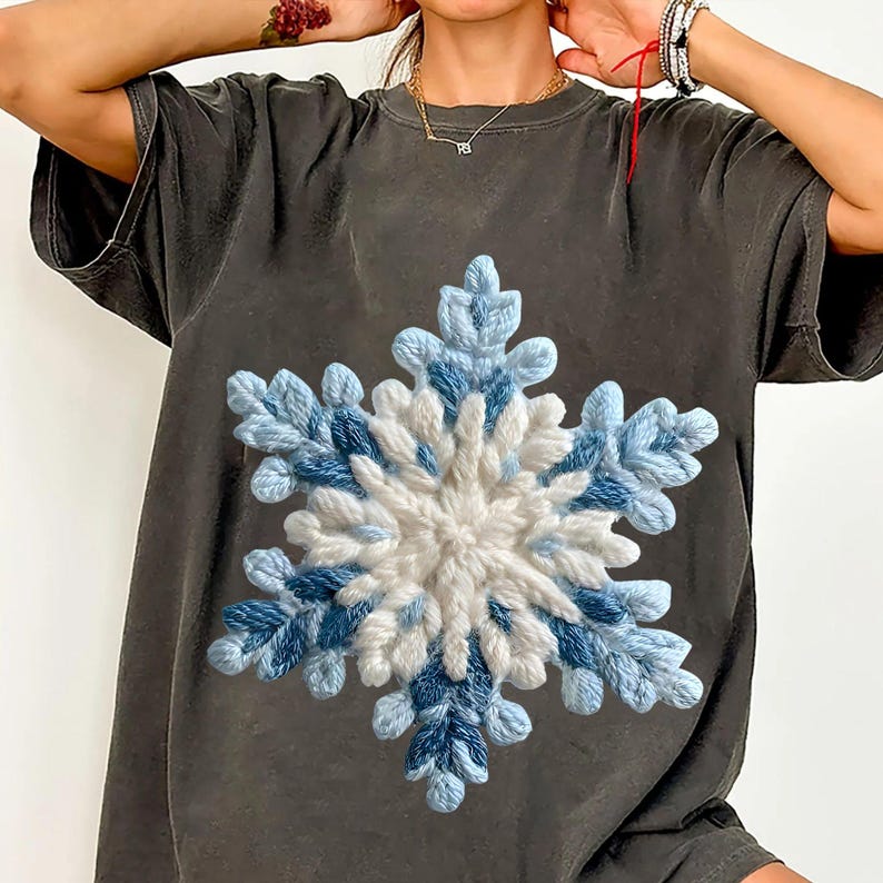 May include: Dark grey t-shirt with a large, textured snowflake design. The snowflake is made with white and blue yarn, creating a 3D effect. The t-shirt has a relaxed fit and short sleeves.