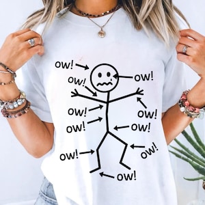 May include: White t-shirt featuring a black stick figure with the word "ow!" pointing to various body parts. The stick figure has a sad face. The shirt is a casual, humorous design.